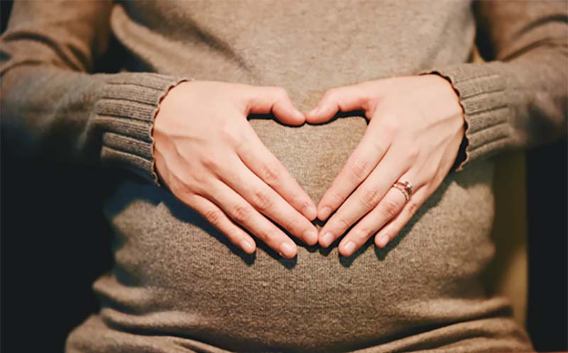 Pregnant mother forming a heart shape with her hands over her stomach representing total care, including shared maternity care of unborn child